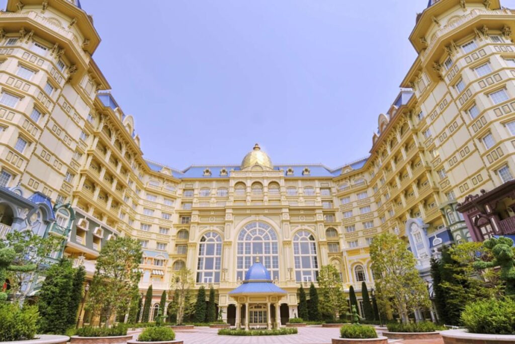 東京ディズニーランドホテルの外観。左右対称の豪華なヴィクトリア朝様式の建築。正面玄関には青いドームの車寄せがあり、周囲には美しく手入れされた植栽やツピアリーが配置されている。青空の下にそびえる夢のようなホテルの全景。