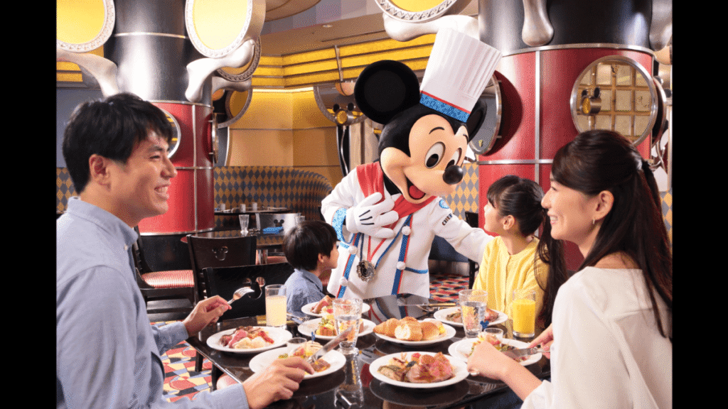 ディズニーアンバサダーホテル内のレストラン「シェフ・ミッキー」にて、朝食ビュッフェを楽しむ家族（父親、母親、娘、息子）のテーブルを訪れた、白いコックコート姿のミッキーマウス。家族全員が笑顔でミッキーを見つめており、テーブルの上にはクロワッサンやジュースなどの料理が並んでいる。