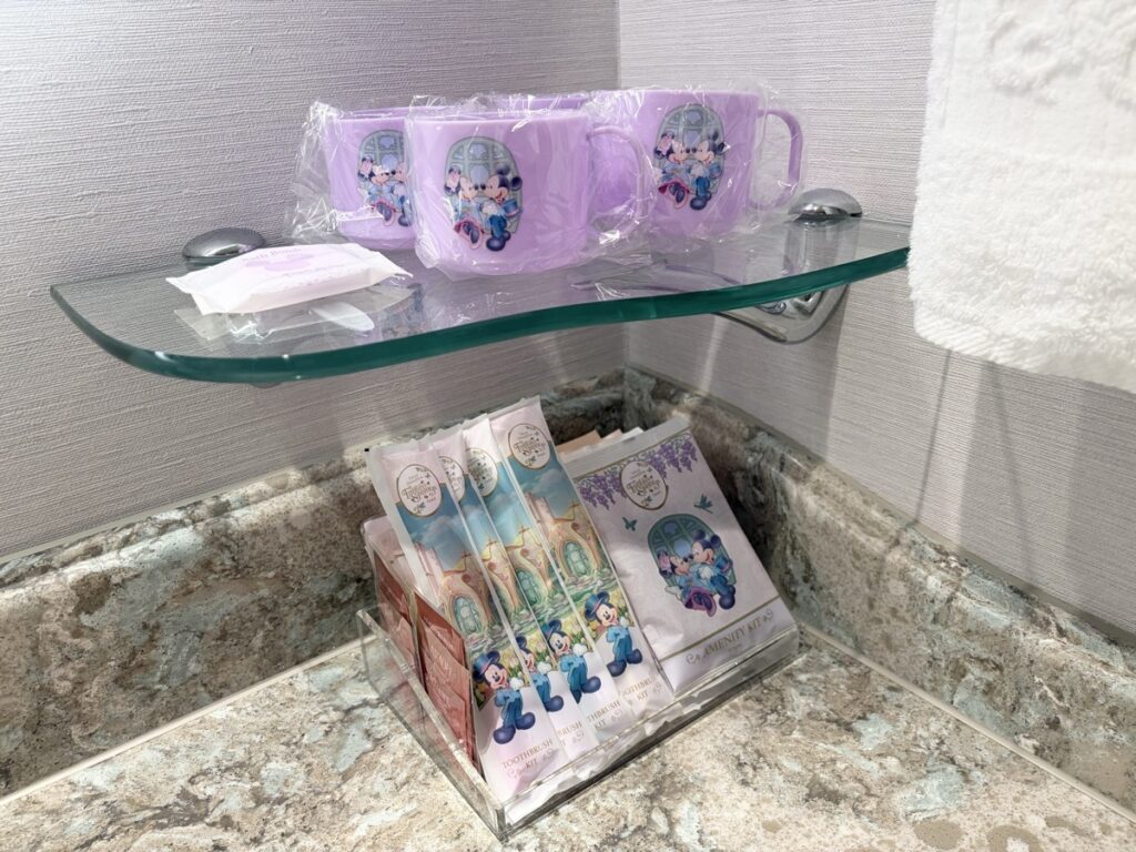 ファンタジースプリングスホテルの客室アメニティ。ガラス棚に紫のミッキー&ミニーデザインのマグカップ2個、下のアクリルスタンドにホテルオリジナルのアメニティキット一式が並ぶ