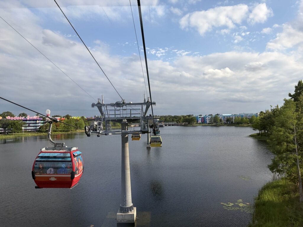 湖の上を進むディズニー・スカイライナーの赤いゴンドラ。晴れた空と対岸の建物を背景に、ワイヤーで運行するロープウェイの様子