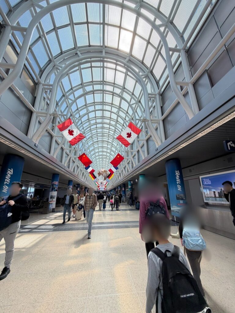 シカゴ・オヘア空港（ORD）のターミナル通路。ガラス張りの天井にカナダ国旗などが並び、乗り継ぎ客が行き交う様子