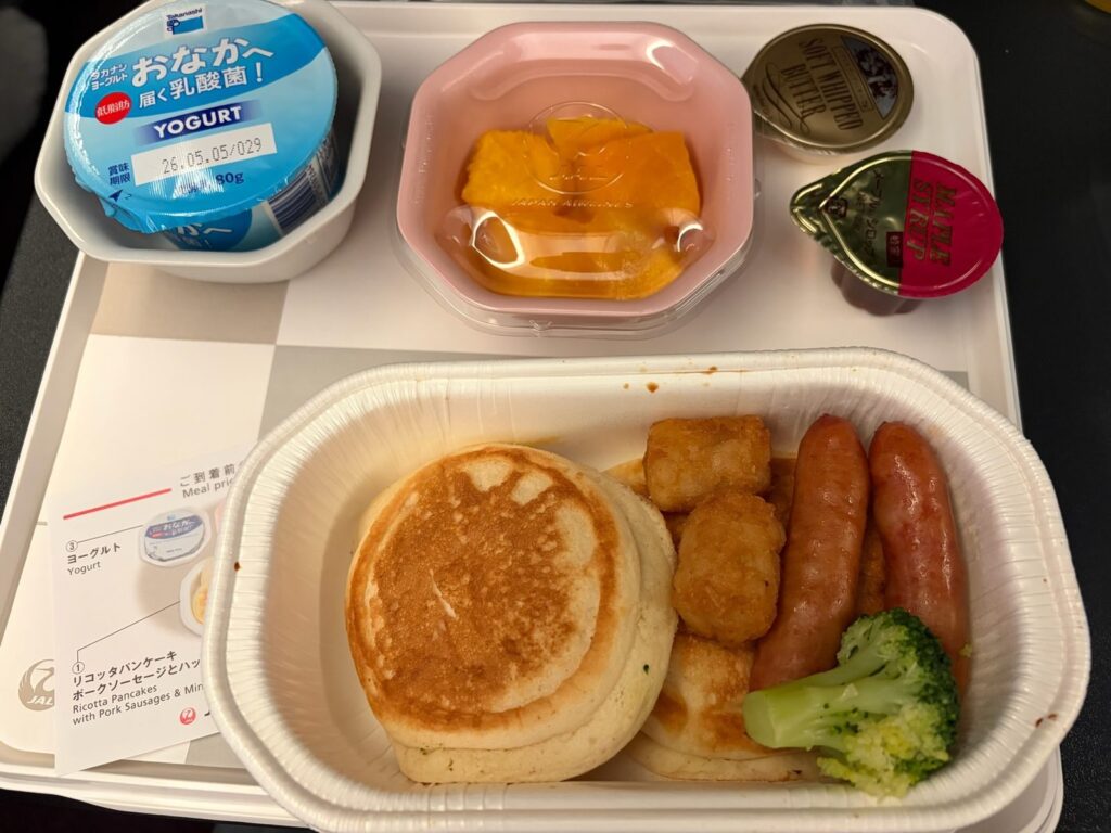 JAL国際線の機内食。リコッタパンケーキ、ポークソーセージ、ハッシュドポテト、ブロッコリー、フルーツ、ヨーグルトが並んだ朝食プレート