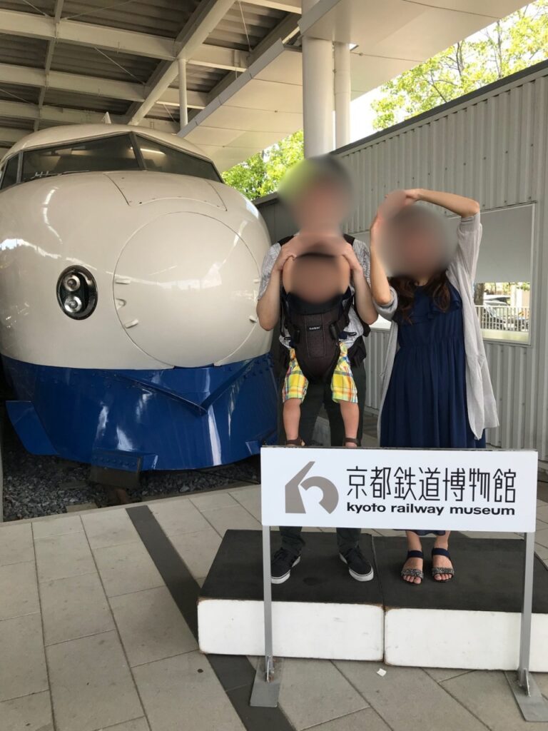 Family posing in front of the round-nosed 0 Series Shinkansen at the Kyoto Railway Museum entrance sign.