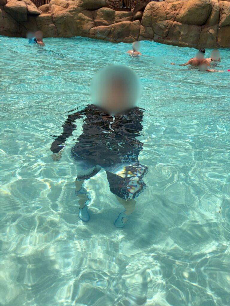 ブリザードビーチの波のプールの浅瀬に立つ、長袖ラッシュガードを着た子ども。奥に岩場が見える
