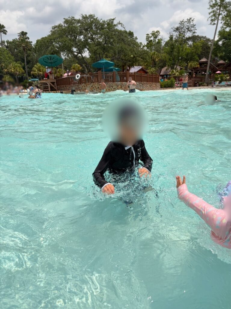 ブリザードビーチの波のプールで、押し寄せた波を体に受けている子ども。奥にビーチとパラソルが並んでいる
