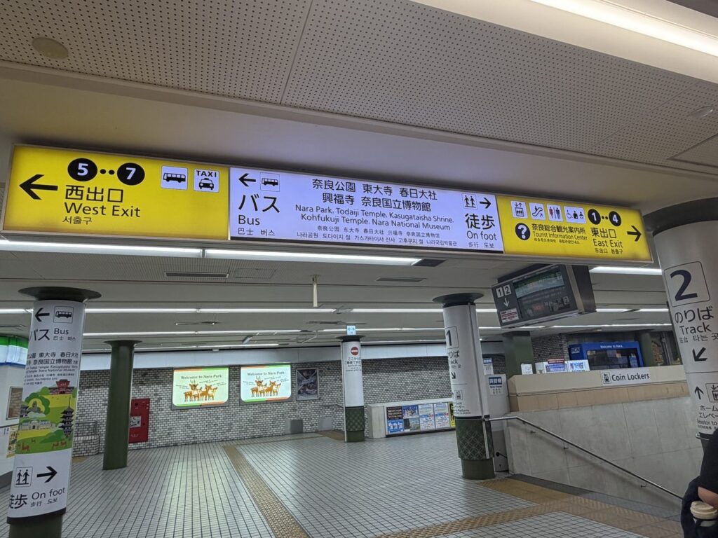 近鉄奈良駅の黄色い吊り下げ案内板。西出口5〜7、バスと徒歩の行き先、東出口1〜4と奈良総合観光案内所の表示が並ぶ